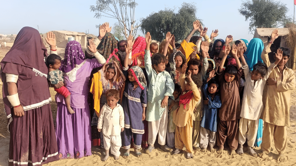 A vibrant gathering of Marwari women and children, dressed in traditional attire, raising their hands high in unison, waving at the camera, under the clear sky of their rural homeland.