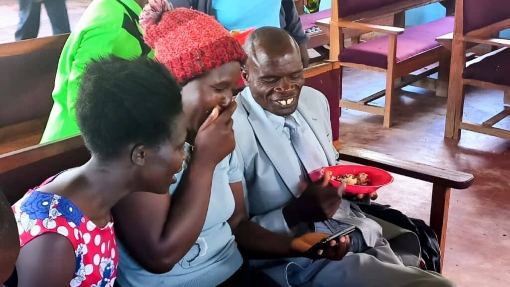 Three people sitting on wooden benches share a joyful moment while looking at a smartphone. The man on the right, dressed in a suit, holds a plate of food.