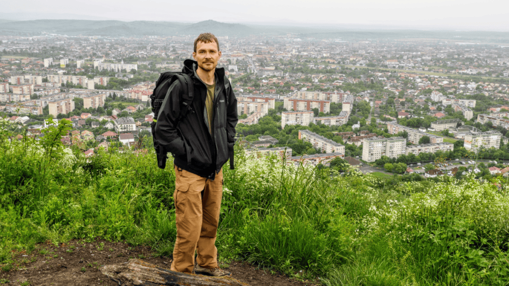 A man wearing a backpack and outdoor gear stands on a grassy hill overlooking a city with numerous buildings and greenery.