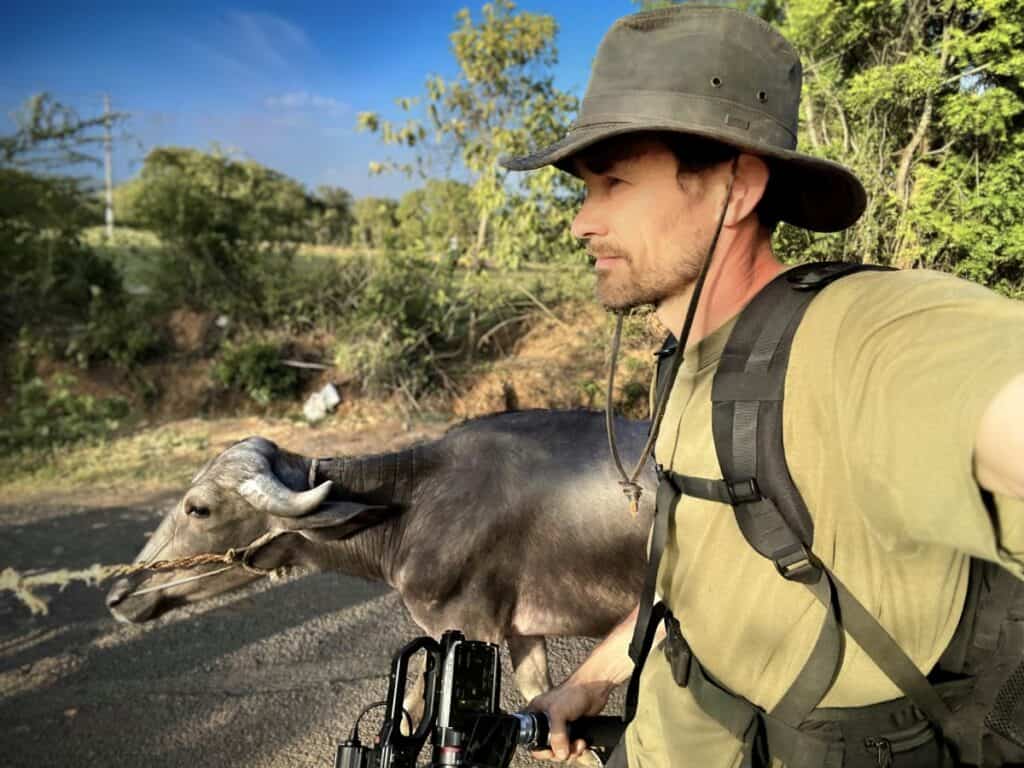 A man in a wide-brimmed hat and backpack walks beside a water buffalo, holding a camera, with greenery in the background.