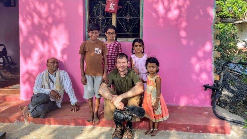 A man crouches in front of a pink house with five smiling children and an older man sitting nearby.