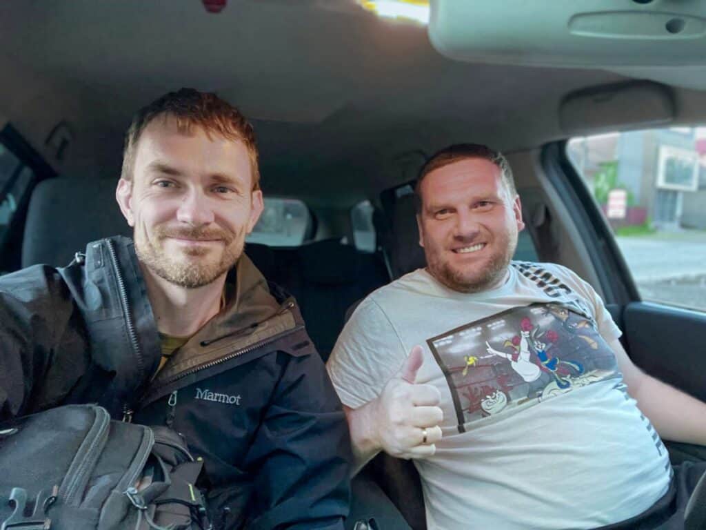 Two men sitting inside a car, smiling at the camera. The man on the right gives a thumbs-up, and the man on the left, Joshua Martyn, holds a backpack.