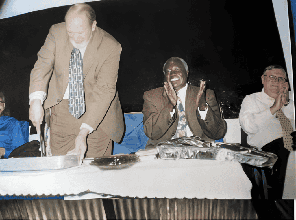 Three men sitting at a table, with one man cutting a cake while others clap and smile.