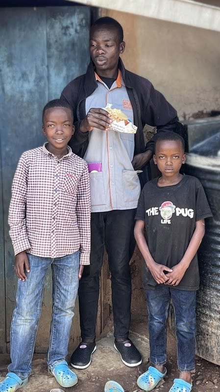 An older boy standing with two younger boys outside a home, all three looking at the camera.