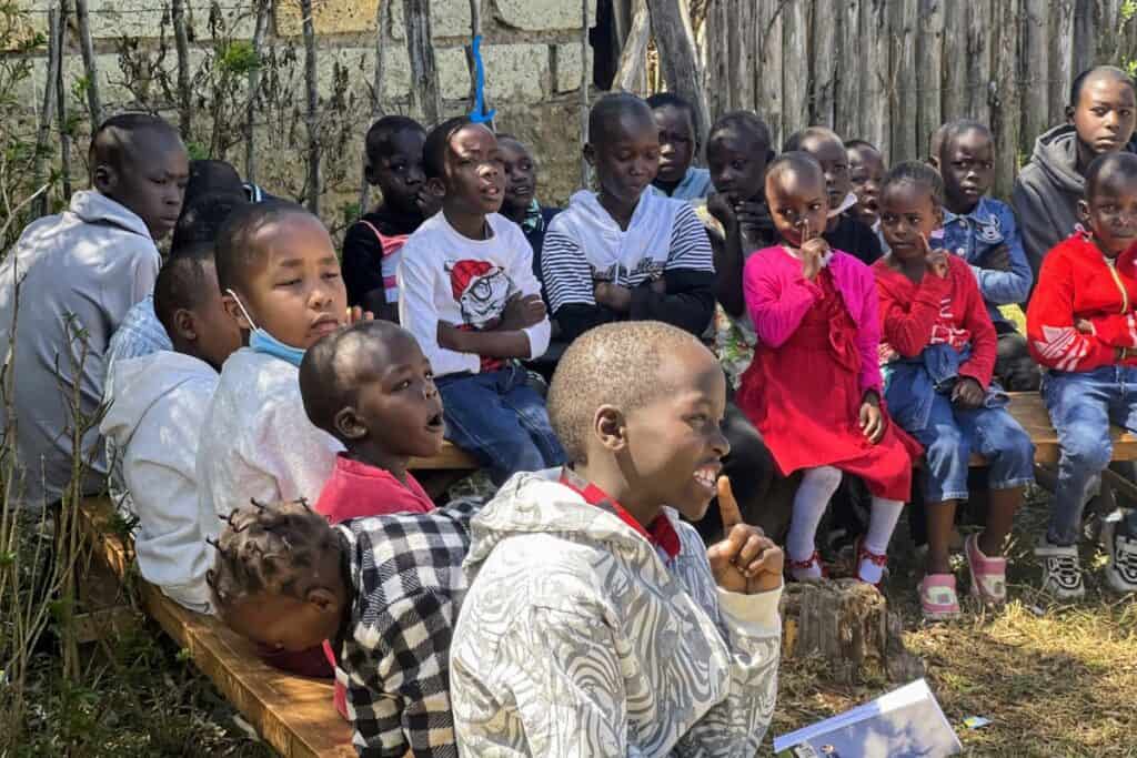 A group of children sitting outside on benches, listening attentively.