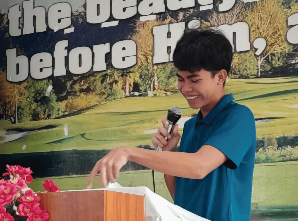 A smiling young man in a blue shirt speaking passionately at a church event, holding a microphone and standing behind a podium decorated with pink flowers.