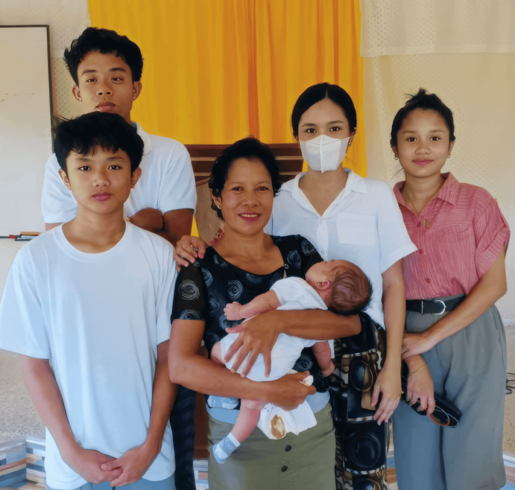 A family of six standing together, with the mother holding a baby. The group includes three children, one wearing a mask, and a warmly lit background featuring a bright yellow curtain.