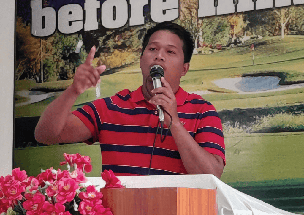 A man in a red and navy striped shirt speaking passionately at a church service. He holds a microphone and gestures with his hand, standing at a podium decorated with pink flowers.