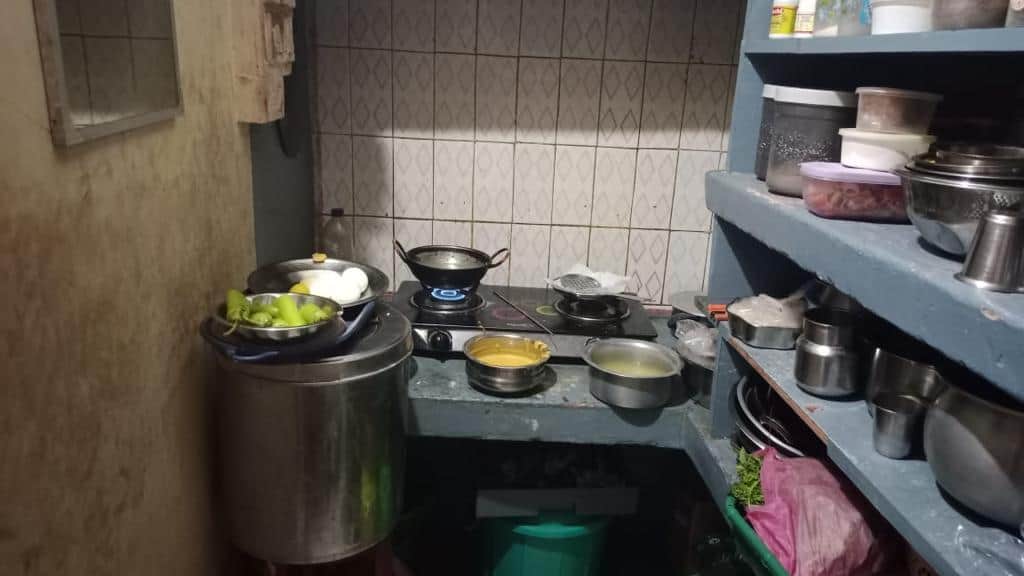 A small, modest kitchen with basic cooking utensils, a stove, and shelves holding various food items and containers.