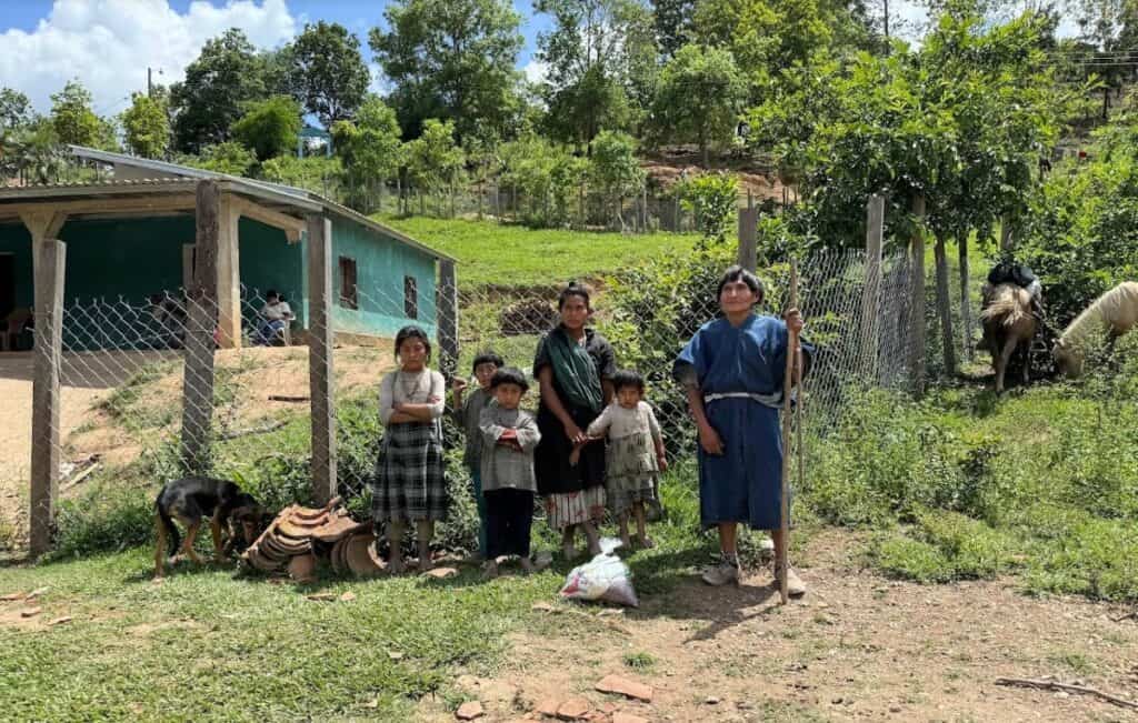 A family of Tolupan Indians stands outside their modest home in the Honduran mountains, surrounded by greenery and animals.