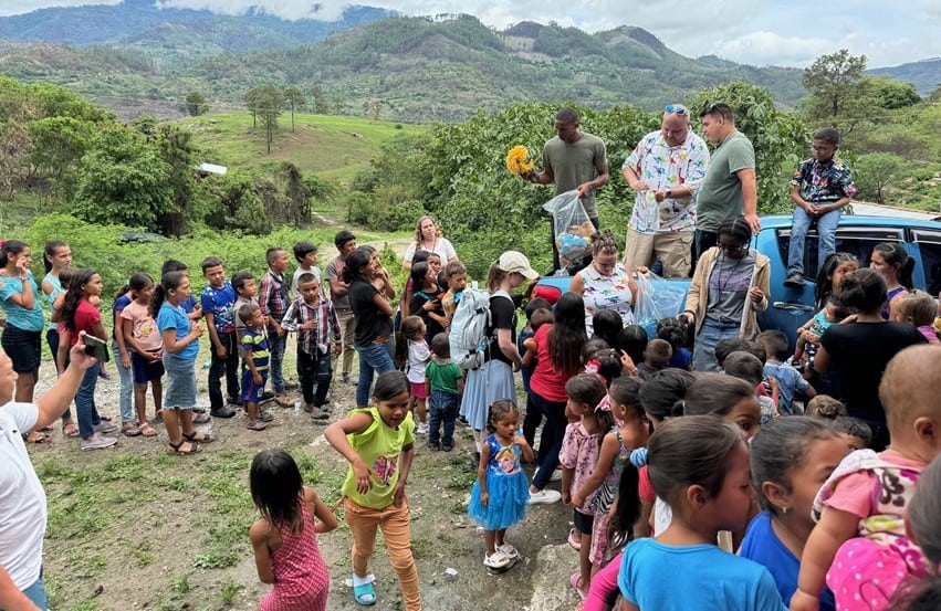 Missionaries hand out toys and supplies to a crowd of children in a rural village, surrounded by scenic Honduran mountains.