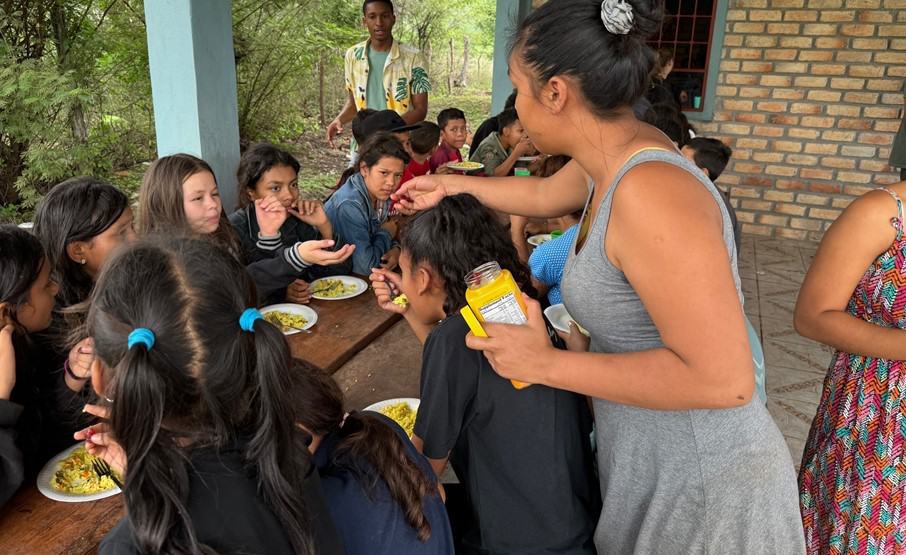 A woman serves children a meal at a feeding center, with several children sitting at a table and enjoying the food.