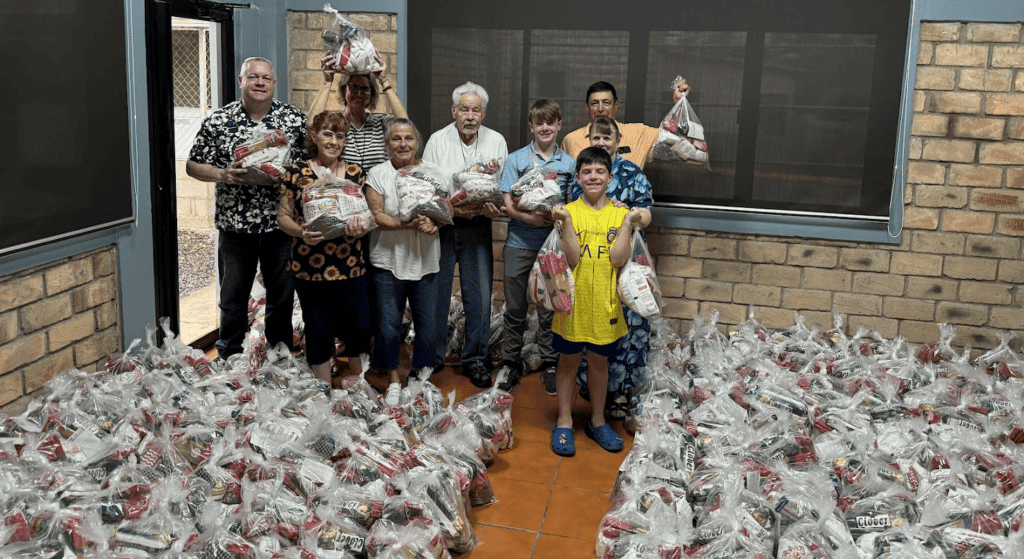 A group of smiling volunteers holds up food bags inside a room filled with hundreds of packaged food bags ready for distribution in Honduras.