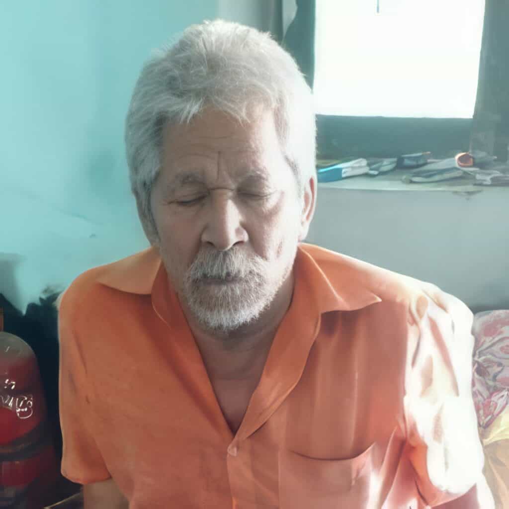 An elderly man with closed eyes, wearing an orange shirt, sits in a modest room with soft lighting. His expression is calm and reflective.