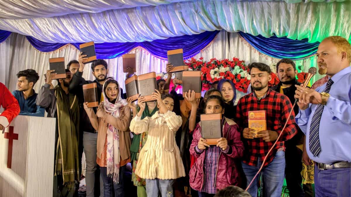 A group of Pakistani Christians, including men, women, and children, pose while holding up Bibles and songbooks they received through the Final Frontiers Foundation's project, standing under colorful decorative drapery at an event.