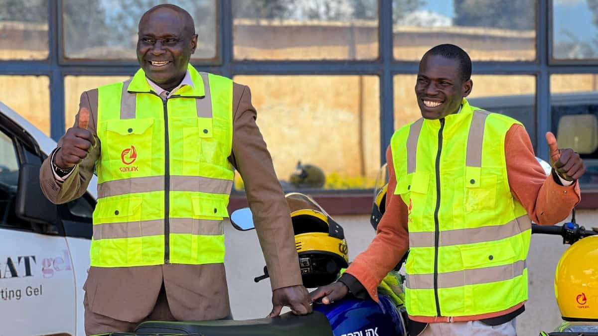 Two joyful pastors wearing high-visibility vests giving thumbs up, one standing next to a motorcycle equipped with various supplies for ministry work.