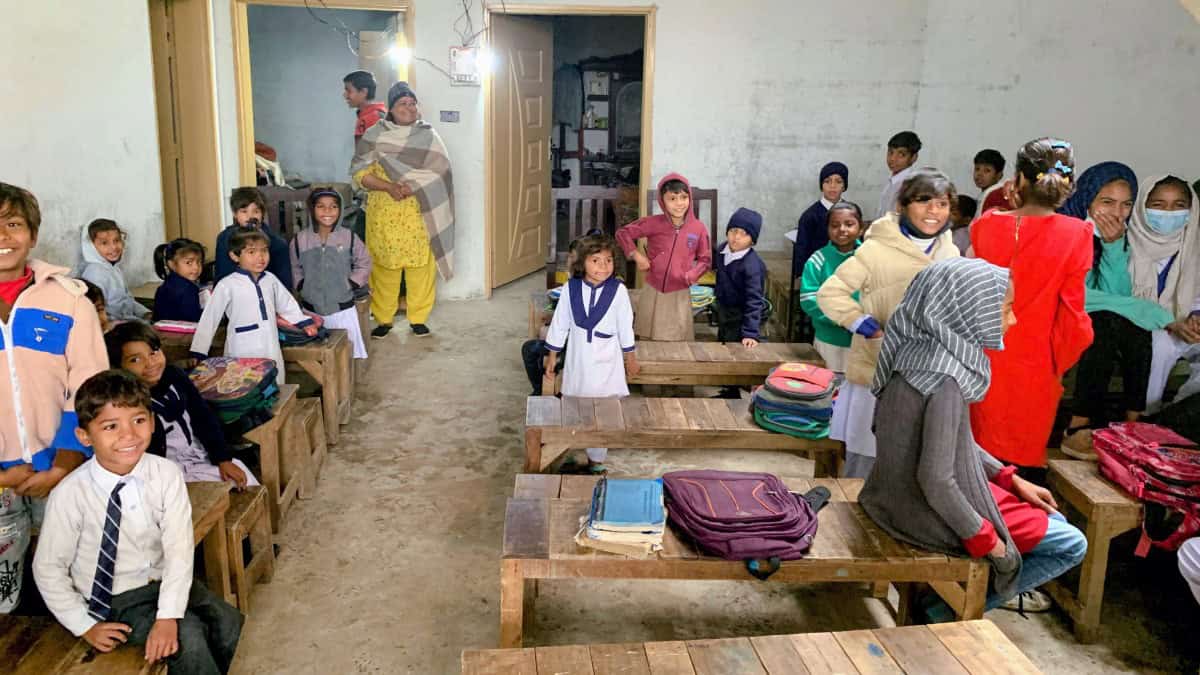 A group of young students, both boys and girls, are sitting on wooden benches in what appears to be a classroom. The students are wearing a mix of colorful clothing including jackets, vests, and traditional attire. They are looking towards the camera with serious expressions. The classroom has a simple, rustic appearance with worn wooden floors and walls. Some backpacks and belongings are visible on the floor near the students.