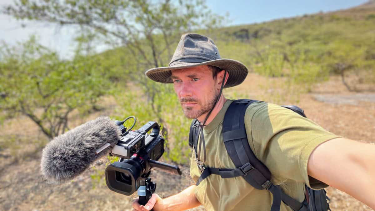Joshua Martin in a wide-brimmed hat and backpack holds a camera with a microphone attached, capturing footage in a dry, scrubby landscape.