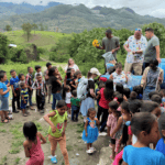 Group of children and adults gathered outdoors in a village in Honduras, with volunteers distributing items from a vehicle.