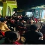 A group of people sitting in chairs at night time in the street. On a nearby building the Jesus film is being projected onto the wall. In the background is a small grocery shop that is lit up.