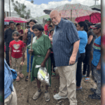 A missionary stands beside an elderly woman from a remote community in Honduras, surrounded by local families holding umbrellas under a cloudy sky.