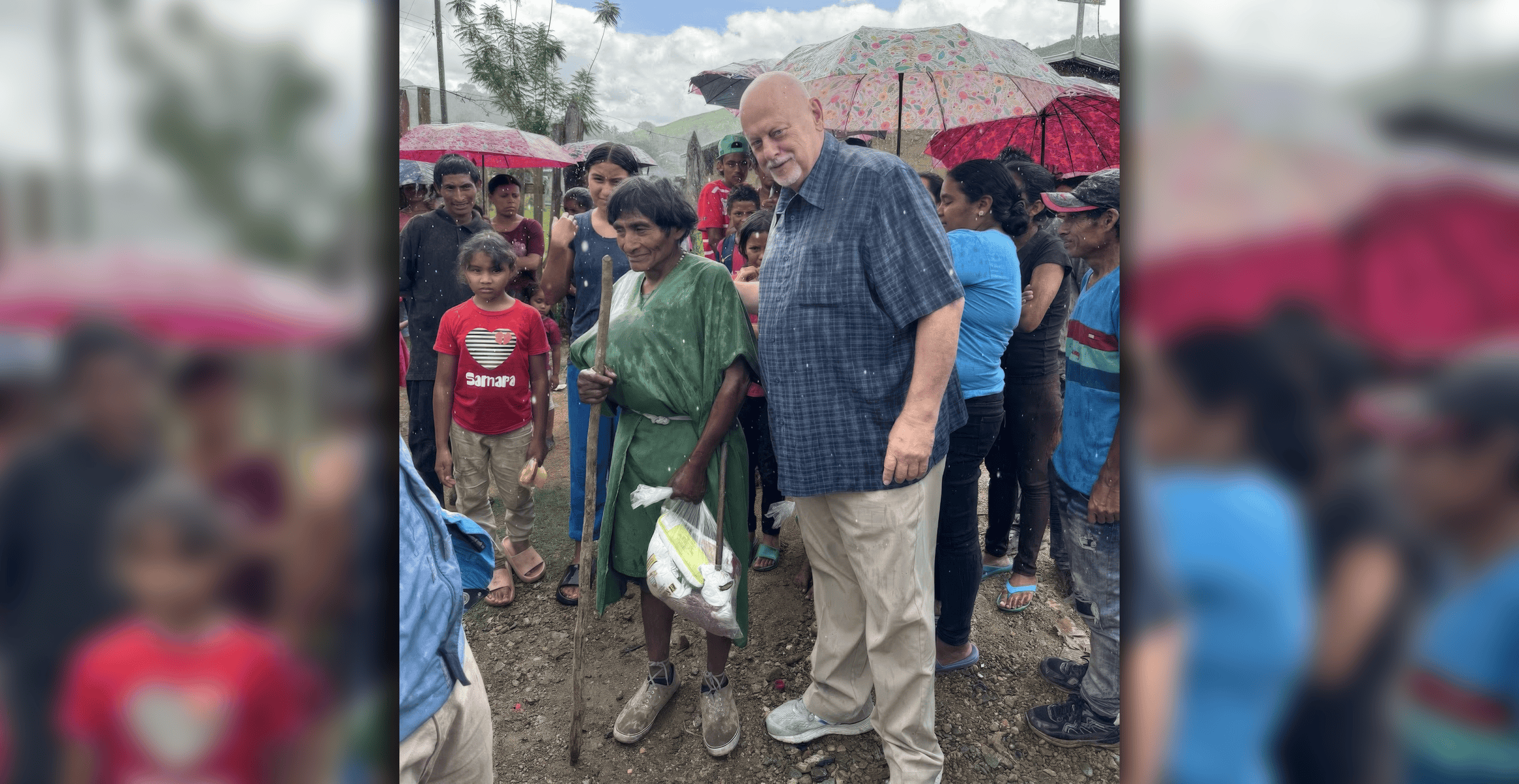 A missionary stands beside an elderly woman from a remote community in Honduras, surrounded by local families holding umbrellas under a cloudy sky.