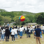 A large group of youths in white shirts enthusiastically playing a volleyball-style game with a giant colorful beach ball in a lush outdoor setting. Facilitators in blue shirts assist and supervise as the group enjoys teamwork and fun amidst a backdrop of trees and hills.