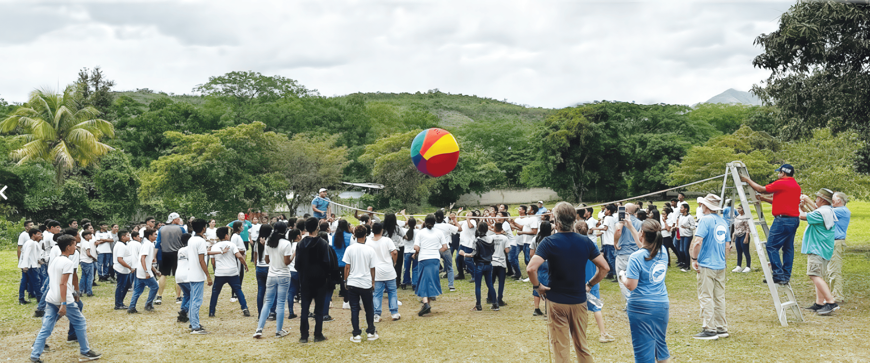 A large group of youths in white shirts enthusiastically playing a volleyball-style game with a giant colorful beach ball in a lush outdoor setting. Facilitators in blue shirts assist and supervise as the group enjoys teamwork and fun amidst a backdrop of trees and hills.