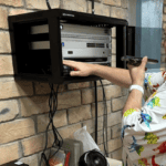 A man wearing a colorful dinosaur-patterned shirt works on installing a Starlink system in a wall-mounted rack at a facility in Honduras.